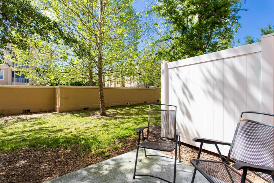Two black metal mesh chairs rest on a shaded concrete slab beside a white privacy fence and green lawn.