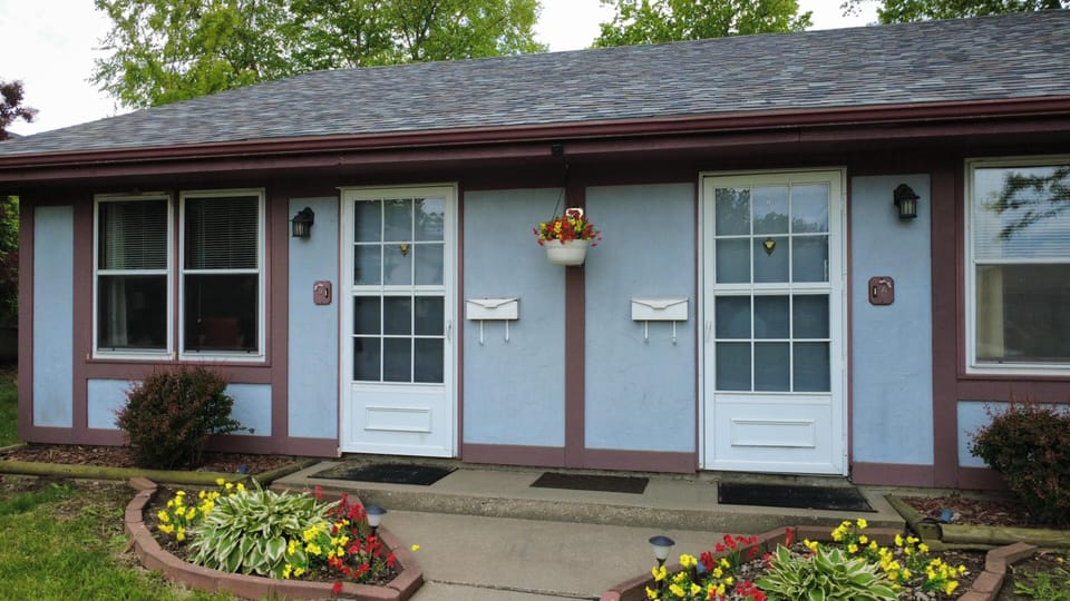 Entrance area with white-framed glass doors, light blue stucco walls, brown trim, and colorful flower beds.