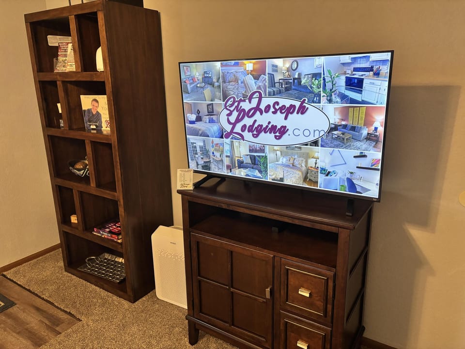 Flat-screen TV sits atop a dark wood cabinet beside a tall, open shelving unit filled with books and decor.