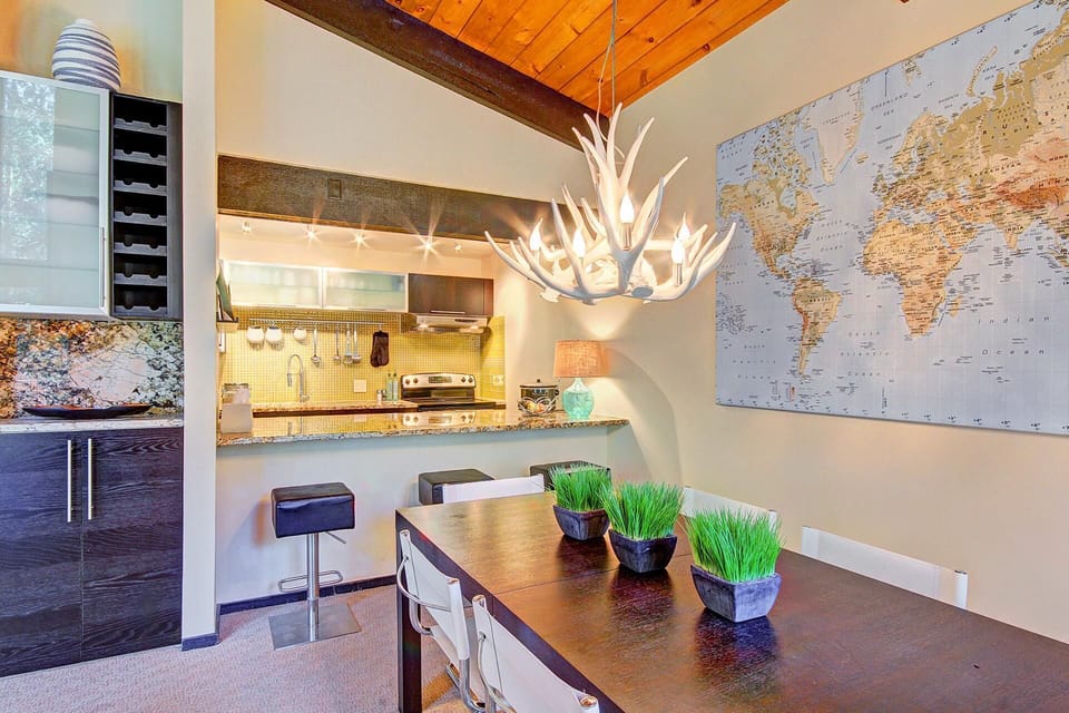 Modern kitchen and dining area with a dark wood table, green plants as a centerpiece, an antler-style chandelier, and a large world map on the wall.