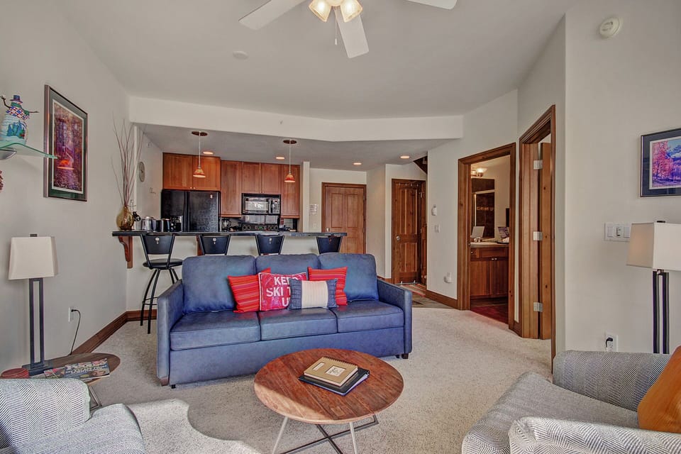A cozy living room with a blue couch, two armchairs, a round wooden coffee table, and an open kitchen with bar stools in the background. A ceiling fan is mounted above, and a hallway is visible.