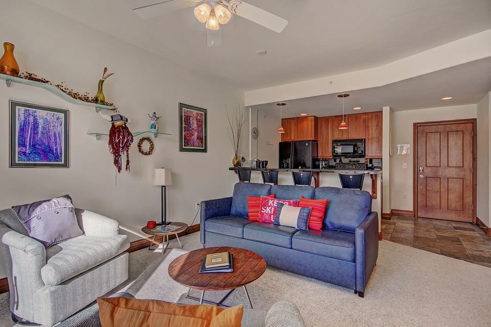 Modern living room with a blue sofa, two chairs, coffee table, wall art, and kitchen in the background. The kitchen features wooden cabinets, a bar counter with stools, and black appliances.