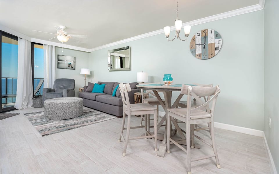 The living area features a gray sofa with queen sleeper, a round coffee table, and a dining set with a beach view
