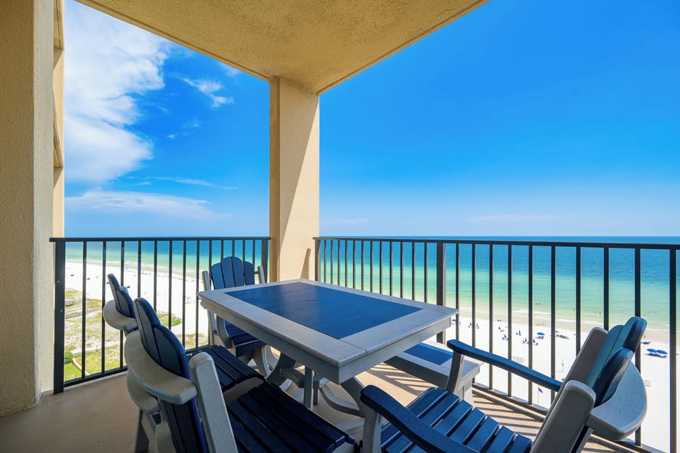 The balcony features an adirondack-style patio set, offering a panoramic view of the beach and clear blue skies