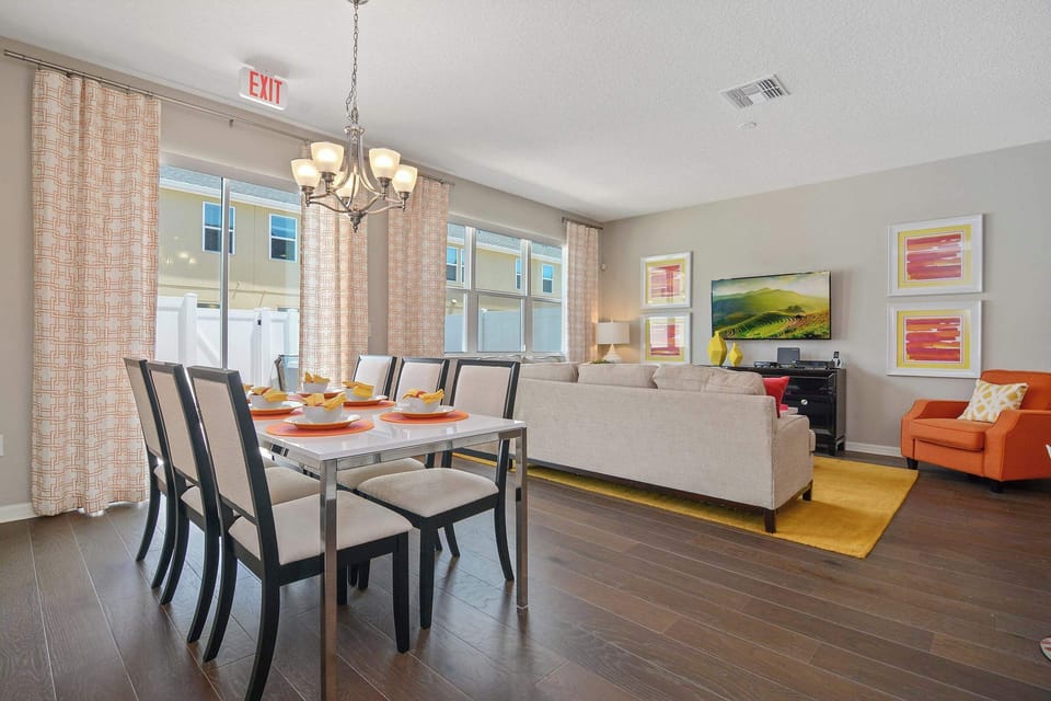 Bright great room with a wall of windows-modern dining set beside a comfy lounge with flat-screen TV; cheerful orange and yellow accents throughout.