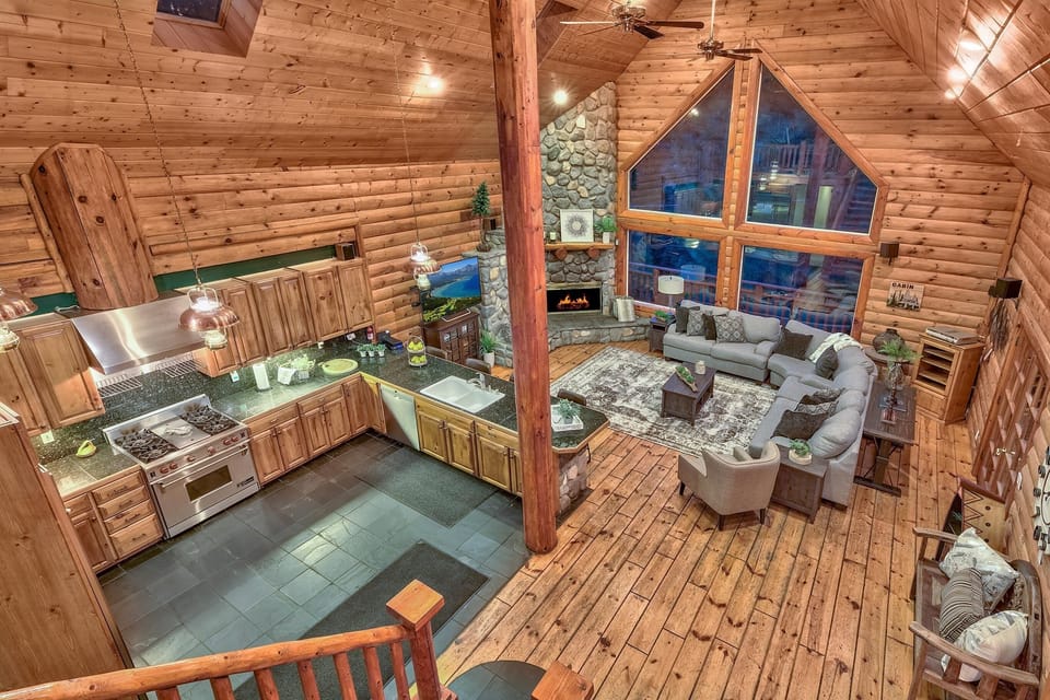 Open-concept living area with stone fireplace, large triangular windows, gray sectional sofa, and wood-paneled kitchen.