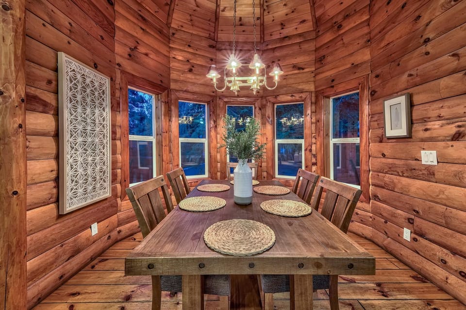 Rustic dining room with six wooden chairs around a thick plank table, surrounded by log cabin walls and multiple windows.