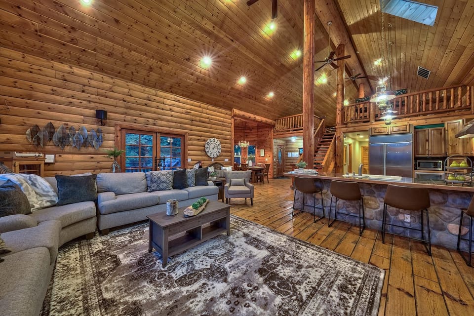 Vaulted knotty pine ceiling and walls rise above a patterned area rug, gray sectional sofa, and stone-front kitchen island with barstools.