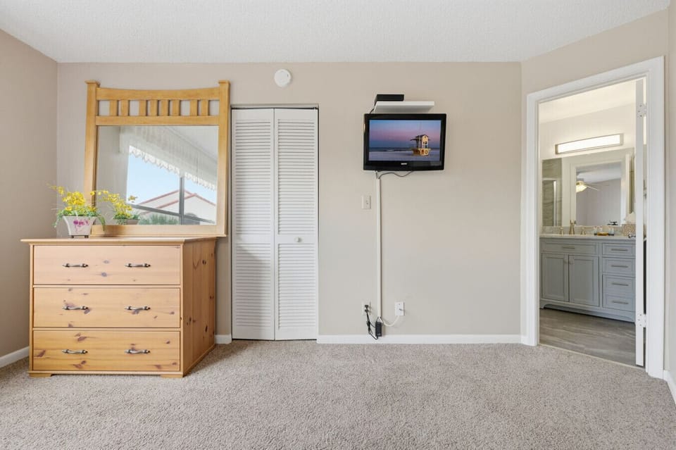 Comfortable vacation rental guest bedroom featuring a flat-screen TV, spacious dresser with mirror, closet storage, soft carpeting, and convenient access to the bathroom. Ideal for a relaxing beach vacation.