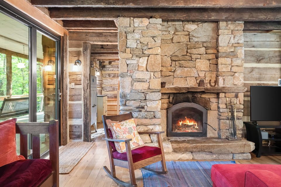 Elegant rustic living room featuring a grand stone fireplace, wood-burning stove, rich antique-style furnishings, and serene forest views through expansive glass doors.