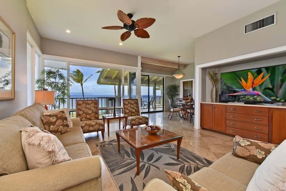 Open-concept living room with tropical furnishings, ceiling fan, and sliding glass doors opening to a covered lanai with ocean views.