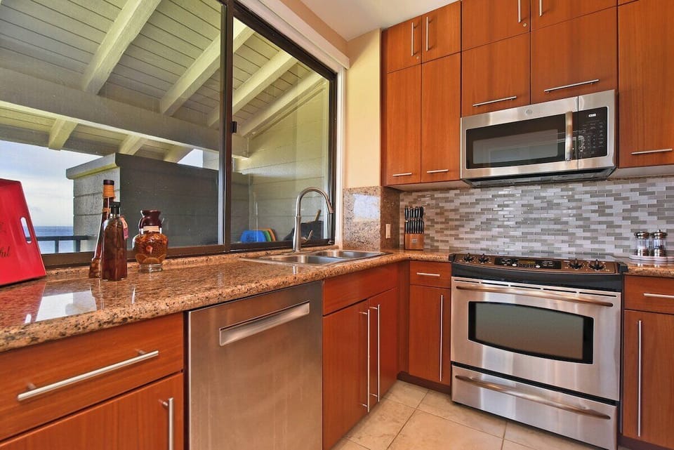 Modern gourmet kitchen with stainless steel appliances, granite countertops, custom wood cabinetry, and a large window opening to the covered lanai with ocean breezes.