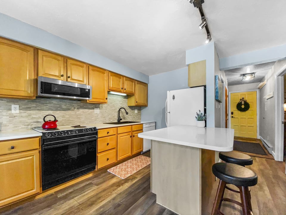 A modern kitchen with wooden cabinets, stainless steel appliances, a white countertop with bar stools, and a yellow door in the background.