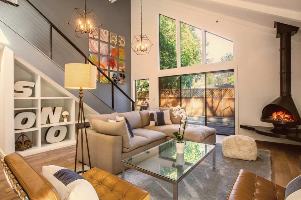 Stylish, sunlit living room with soaring ceilings, large windows, and a cozy modern fireplace. Relax on the plush sectional, enjoy chic décor, and step out to a serene private deck—perfect for peaceful mornings or evening lounging.