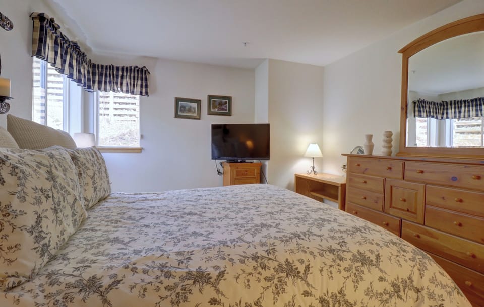 A bedroom with a floral-patterned bedspread, wooden furniture, a dresser with a TV on it, two pictures on the wall, and windows with striped curtains.
