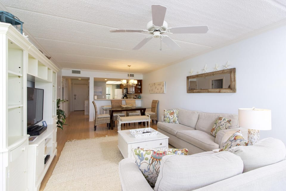 Open living and dining area with beige sofas, coastal-patterned pillows, white entertainment center, and hardwood floors.