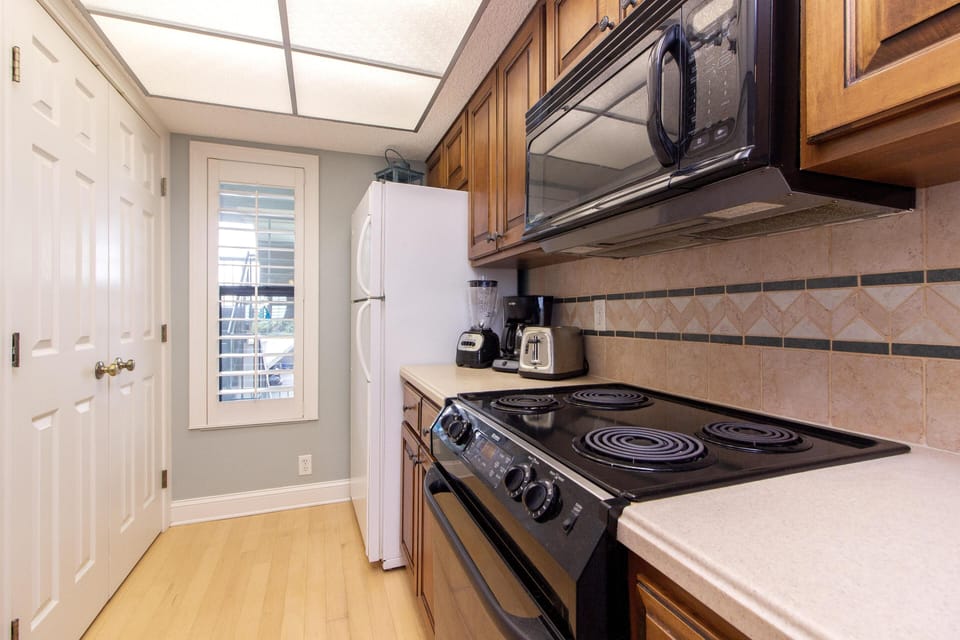 Galley kitchen with black electric stovetop, microwave, wood cabinets, tiled backsplash, and light wood flooring.
