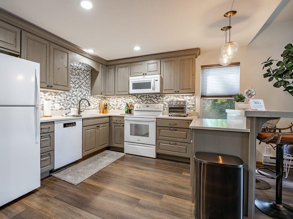 Gray wood cabinets and mosaic tile backsplash surround white appliances in the kitchen with wood-look flooring.