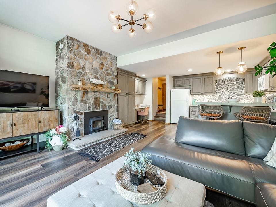 The living room features a stone fireplace, gray leather sofa, tufted ottoman, and a wood console under the TV.
