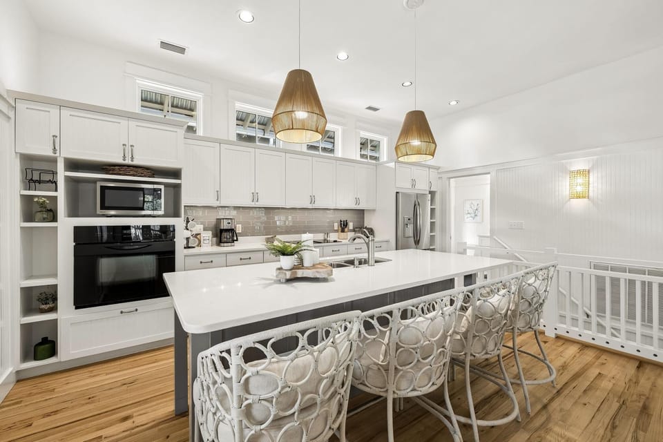 White kitchen with beadboard cabinetry, quartz island, rattan pendant lights, and woven-backed bar stools.