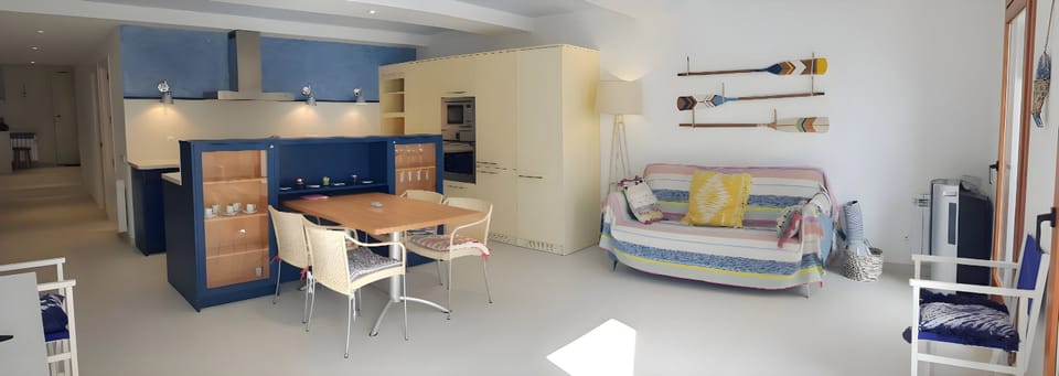 The open-plan living space features a dining table with four chairs, positioned near a fully equipped kitchen with a blue cabinet and glass display shelves.  A comfortable couch with colorful pillows and a blanket adds a touch of vibrancy to the room.