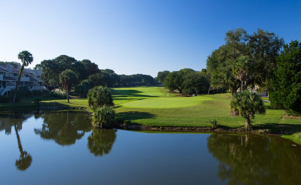Championship golf course stretches across manicured greens, featuring scenic water hazards and tropical landscaping under clear blue skies.
