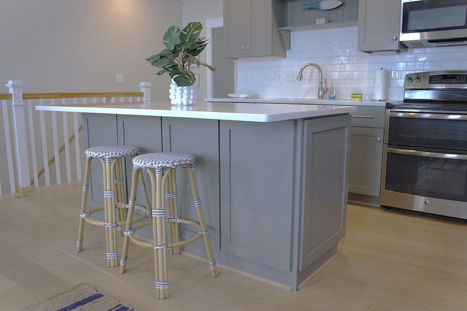 The island provides ample workspace and includes two stylish rattan barstools with woven seats. Stainless steel appliances, including a refrigerator, oven, and microwave, contribute to the sleek look.