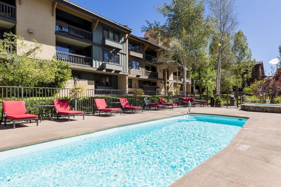 Outdoor swimming pool area with red lounge chairs and multi-story residential buildings in the background. Trees and greenery surround the area.