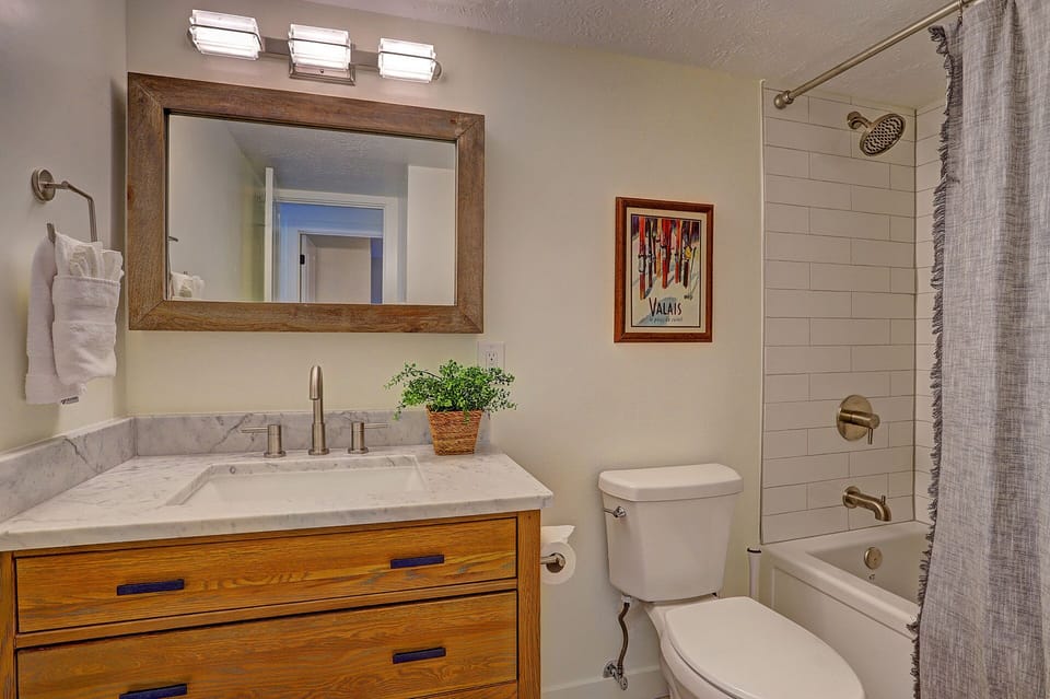 A modern bathroom features a white vanity with a marble countertop, a large rectangular mirror, wall-mounted lights, a toilet, a bathtub with a shower, and a framed picture on the wall.