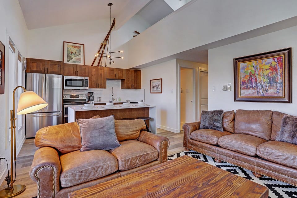 A cozy living room with brown leather couches, a wooden coffee table, patterned rug, modern kitchen in the background, a staircase leading to an upper level, and colorful wall art.