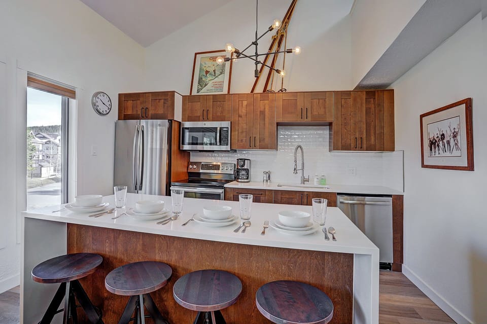 A modern kitchen with wooden cabinets, a white island with four stools, set for dining. Stainless steel appliances, a contemporary light fixture, and framed artwork are visible.