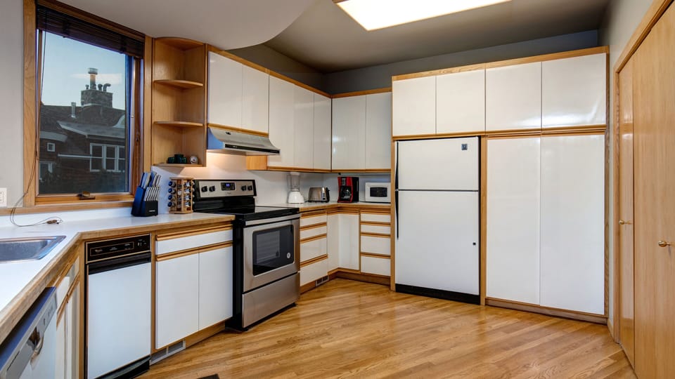 A modern kitchen with white cabinets, stainless steel appliances, a built-in oven, a refrigerator, a dishwasher, a microwave, and wooden flooring. A window offers an outside view.