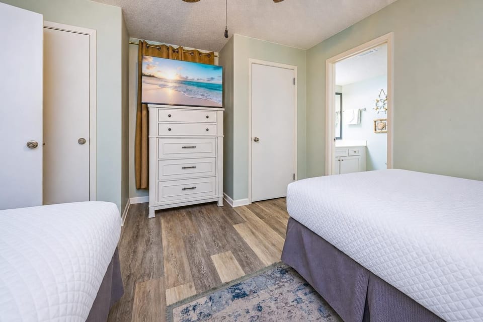 The guest bedroom featuring two double beds, a white dresser with a TV, and access to a bathroom through a nearby door