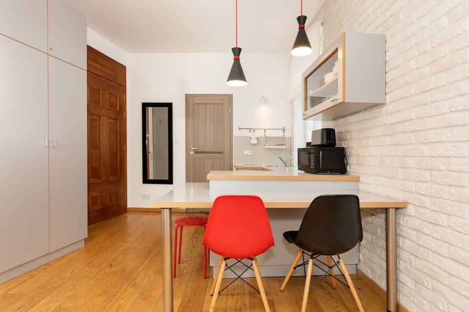 The kitchen area has a white backsplash, grey cabinets, and a dining space with a small table and red stools. Large windows bring in natural light, making the room feel bright and airy.