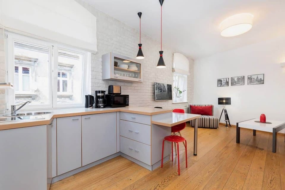This living space has an open-plan layout with a kitchenette visible in the background. The red sofa complements the red bar stools at the kitchen counter, tying the room together with pops of color.