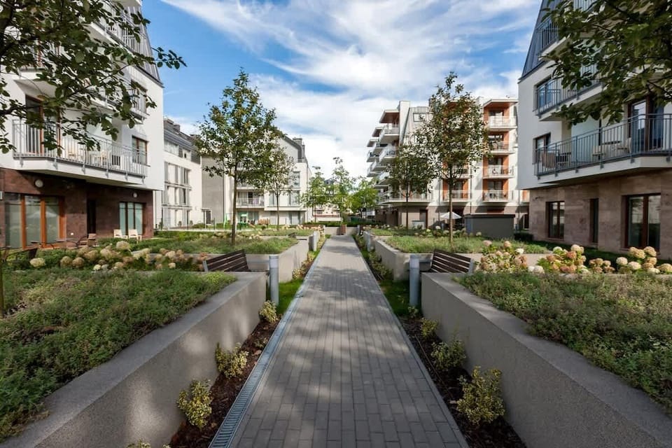A wide outdoor view showing a modern residential complex with well-maintained gardens and paved walkways, surrounded by lush greenery.