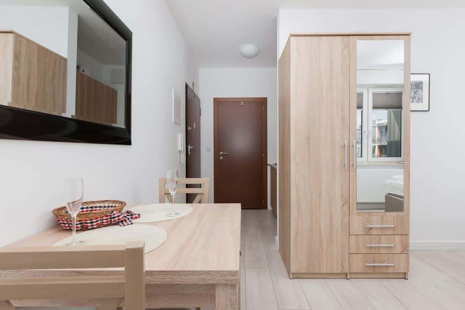  A bedroom or small living area with light wood furniture, including a wardrobe and a television on the wall. The minimalist decor creates a clean and functional vibe.