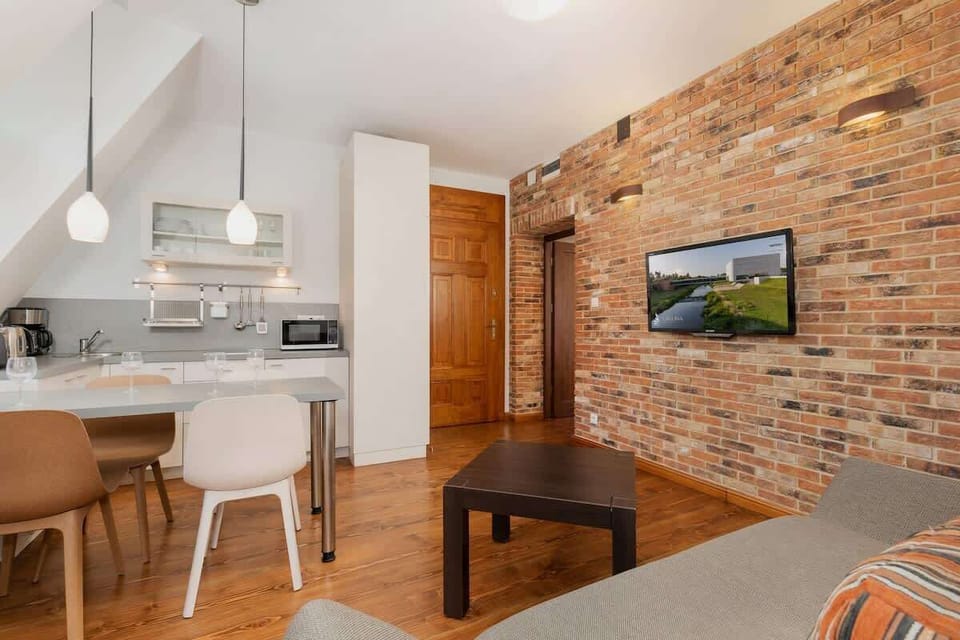  A brick accent wall adds warmth to this open-plan living and dining area. There is a flat-screen TV mounted on the wall, a gray sofa, and a small dining table.