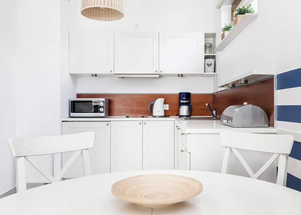 
In the foreground, a side table with a decorative bowl can be seen, and behind it is the kitchen area with white furniture, microwave, coffee machine and essential utensils.