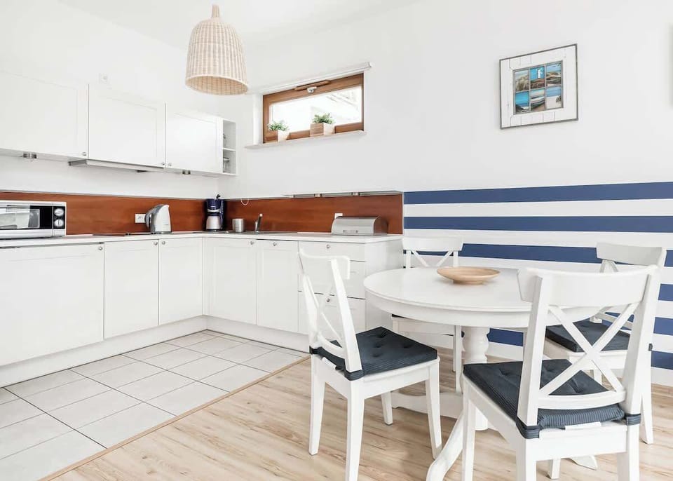 The kitchen is brightly coloured, with white cabinets and wooden accents. There is a dining table with four chairs against the wall with a window. A decorative blue stripe on the wall adds to the charm of the interior.