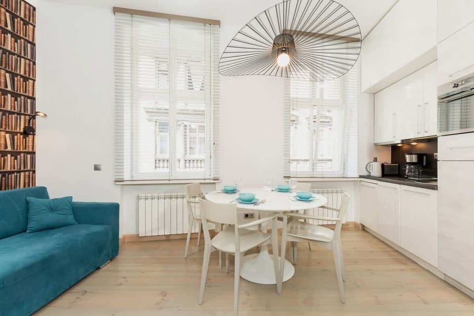 A stylish living room with a teal sofa and white dining table. The kitchen area includes sleek white cabinets and modern appliances, creating an open and inviting space.