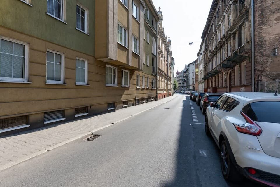 A narrow, European street lined with historic buildings featuring decorative façades. The street is flanked by parked cars on both sides, with tall, charming structures that give the area a classic, old-world atmosphere.