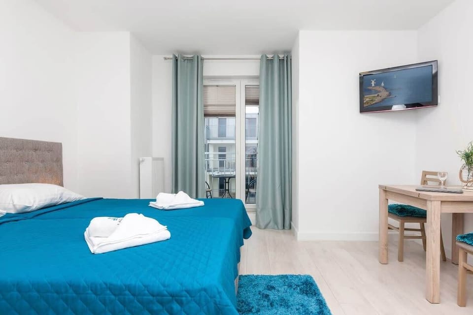 A bedroom showcasing the same queen-sized bed with a blue bedspread, this time with a view of the bed’s foot and side. A plant on a small side table adds a touch of decor, and curtains cover the window.