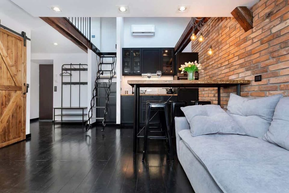A comfortable living room with a gray sofa, with a kitchenette visible in the background. The open-plan design connects the two spaces seamlessly, making it ideal for socializing.