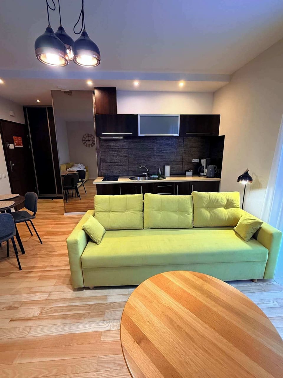 A bright living area featuring a green sofa, a round wooden coffee table, light wooden flooring, and modern ceiling lighting that adds warmth to the space.