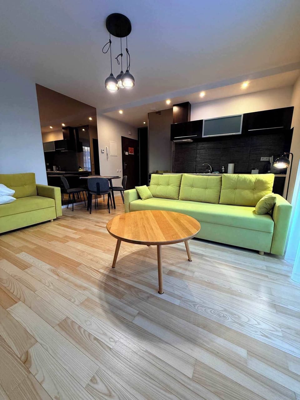 A cozy open-plan interior with a green sofa in the foreground, a dining area behind it, and a dark-toned kitchen accented by stylish pendant lights.