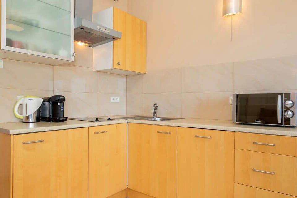 A close-up of a small kitchen with light wood cabinets and appliances like a microwave and kettle. Clean and functional setup.