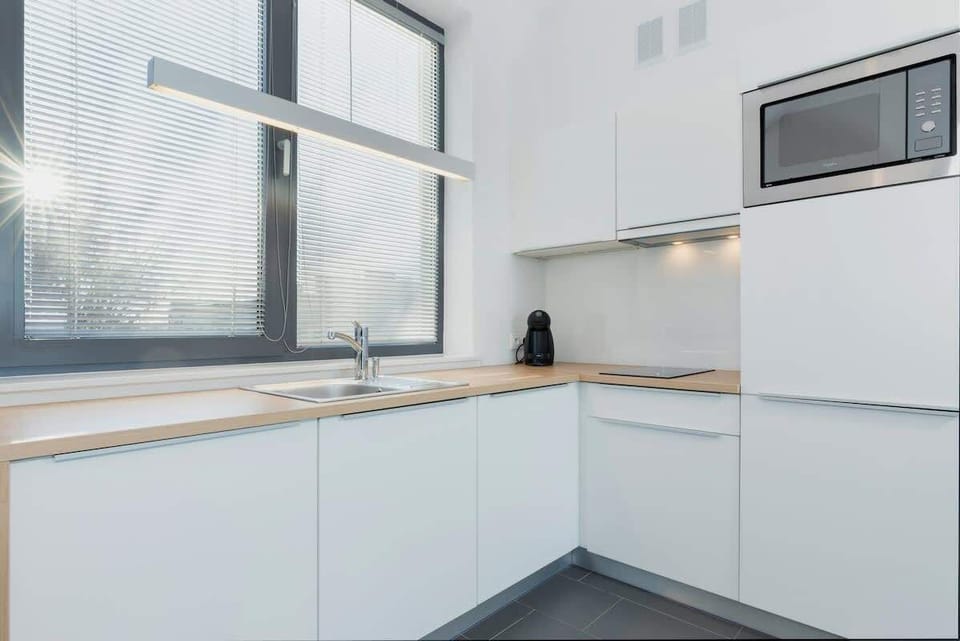 A sleek, modern kitchen with white countertops and cabinets. The large window brings in plenty of natural light and offers a glimpse of the sea outside.