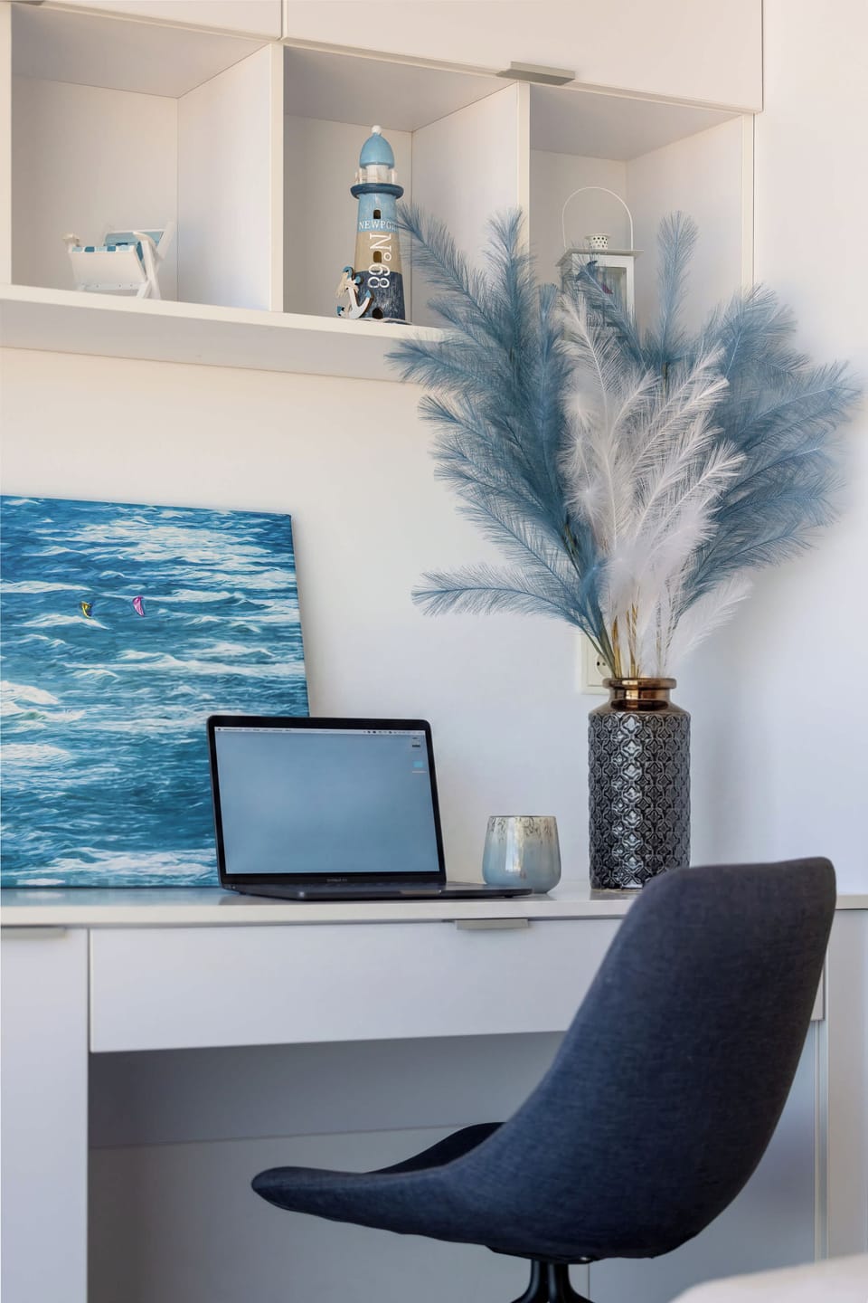 A minimalist white desk with a dark blue swivel office chair. A framed piece of blue abstract artwork hangs on the wall, reinforcing the apartment's coastal design theme.