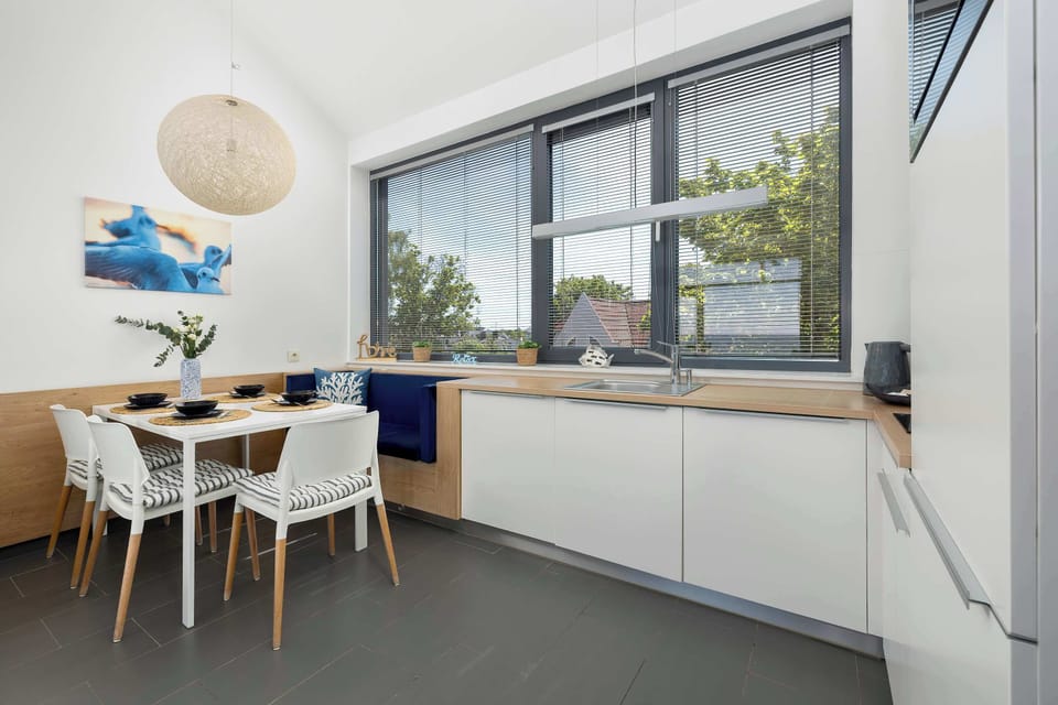 A dedicated dining table set for four is paired with white chairs, positioned adjacent to a long white kitchen counter line. A large window with privacy blinds ensures the space receives plenty of natural daylight.